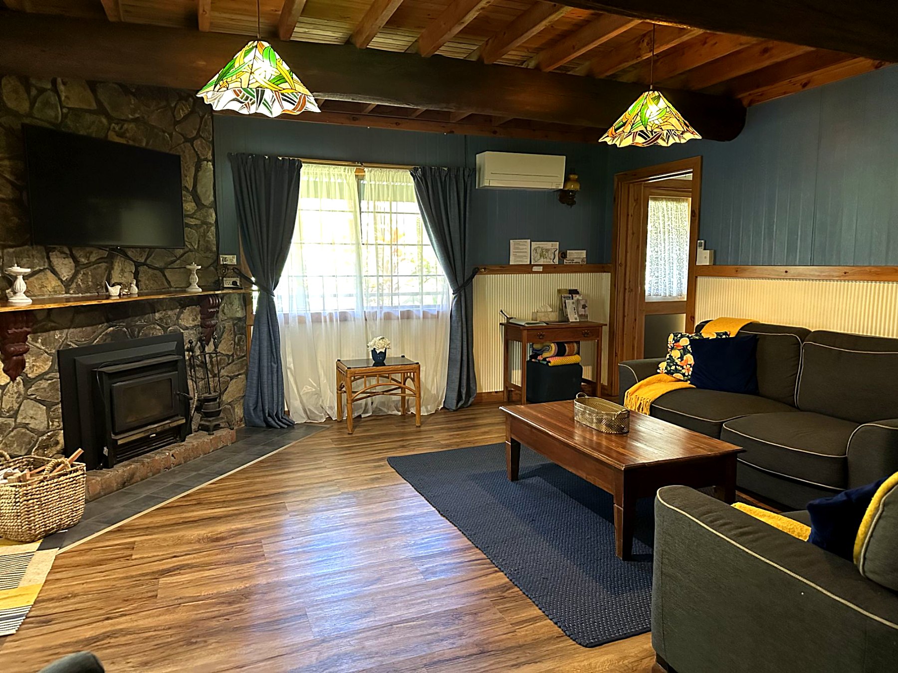 Living room with charcoal sofas, a stone fireplace with a mounted TV, and timber ceiling beams