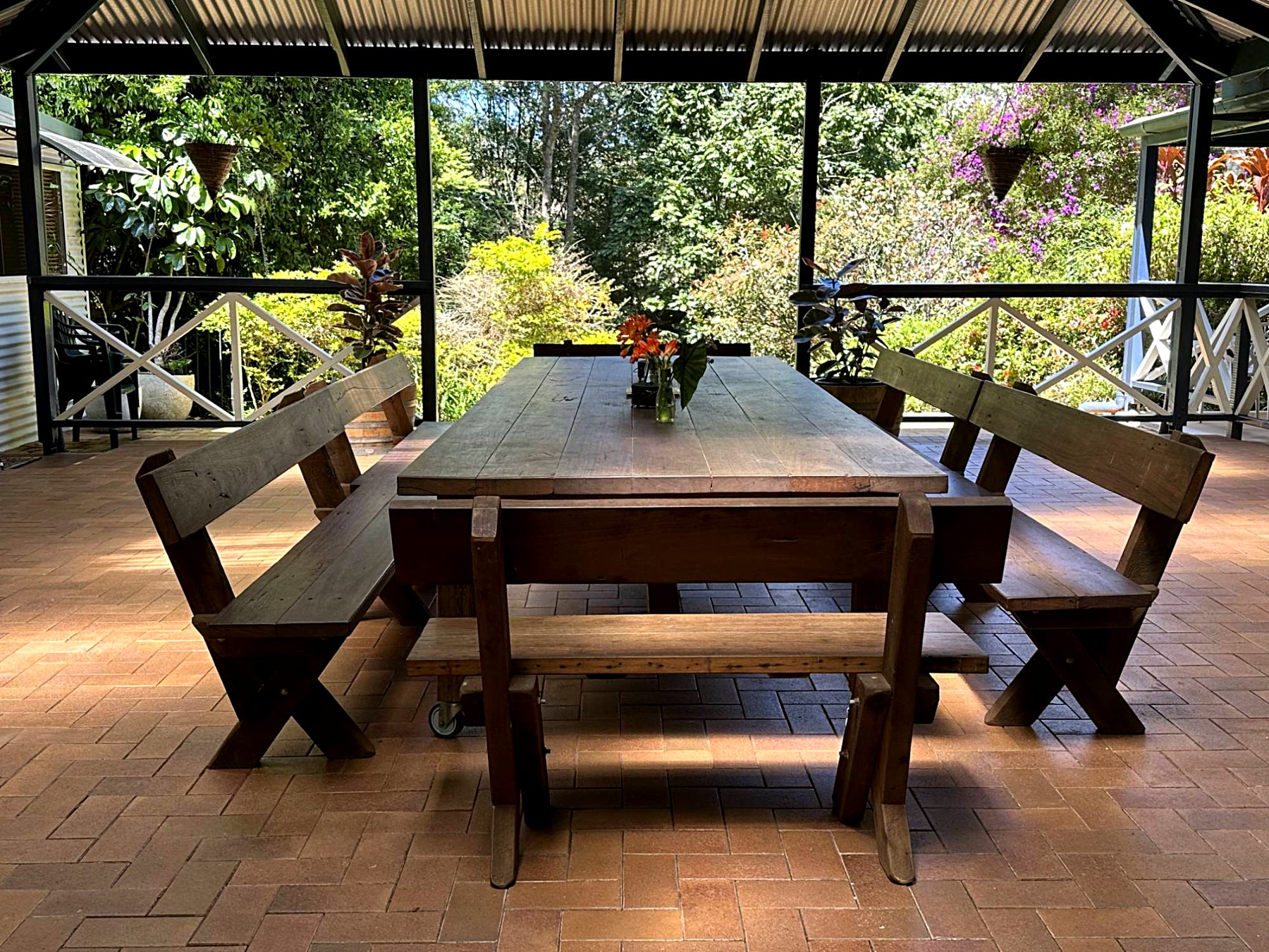 A long reclaimed-timber table with bench seating, under a screened outdoor patio looking onto the garden